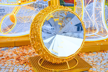 Christmas decor with multifaceted mirrored center. Golden spherical decoration of highly reflective, faceted center sits on stand outdoors near fallen leaves. Light installation glow in backgroundの写真素材