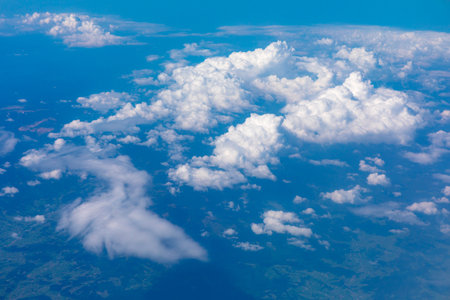 Billowing cumulus clouds drift above a patchwork of green terrain, casting soft shadows over the...