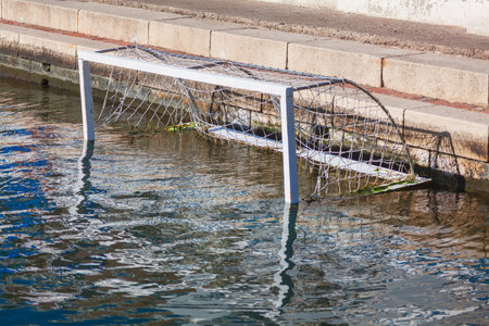 Water polo goal partially submerged in a sunlit body of water. Positioned against a stone ledge, the...