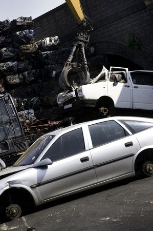 A car scrap yard with a crane lifting a van, piles of crushed cars, and uncrushed cars.