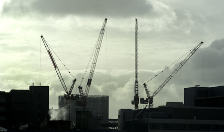Four building cranes silhouetted against sky with city buildingsの写真素材
