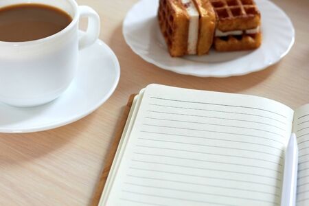 Side view: a cup of fresh coffee, waffles with cream filling on a plate, a notebook are on the tableの写真素材