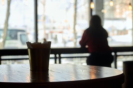 A cylindrical glass napkin on a brown table in a cafe. On a blurred background, the silhouette of a lonely girl at the bar counter. Inside view of the street. Man looking out the windowの写真素材