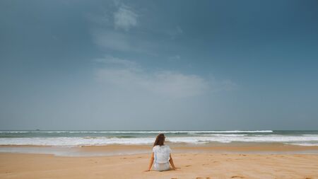 Portrait from the back of a girl sitting on the beach and looking at the oceanの写真素材