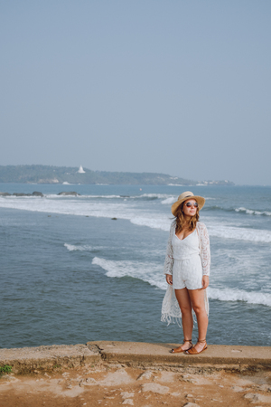 Girl in a hat and sunglasses stands near the ocean on a background of wavesの写真素材