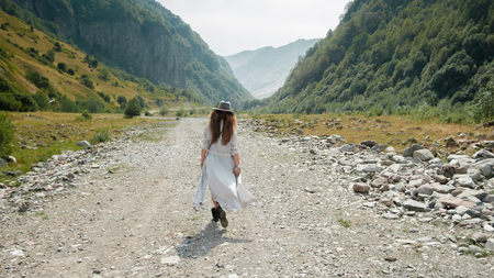 A girl in a hat walks along the middle of the road towards the mountains in the valley in the mountainous country of Georgiaの写真素材