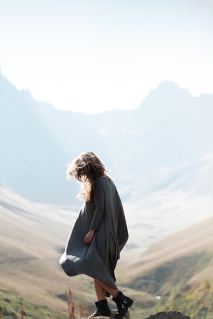Young beautiful girl in a long dress is standing by the cliff in the background of mountains....