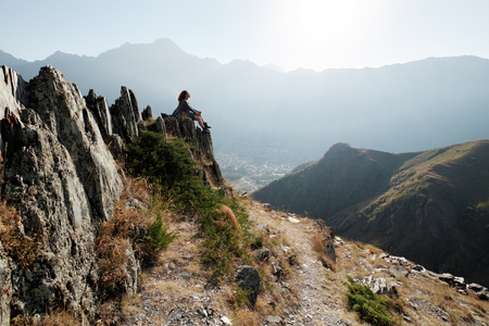 A beautiful girl in a flying dress and hat sits on a cliff overlooking the mountains. Military-Georgian roadの写真素材