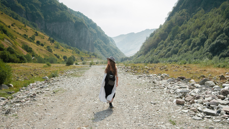 A girl in a hat walks along the middle of the road towards the mountains in the valley in the mountainous country of Georgiaの写真素材