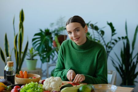 A young girl in a green sweater slices a knife with a knife on a wooden cutting board to make a salad. The concept of healthy proper nutrition, diet. Veganism and Vegetarianism.の写真素材