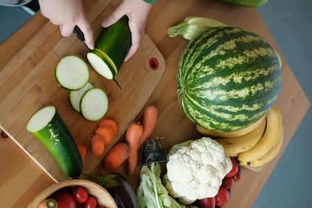 A vegan girl in a green sweater cuts with a knife fresh vegetables and fruits. Fresh healthy vegan and vegetarian food on a wooden table. Slow food, comfortable food, healthy food, clean foodの写真素材