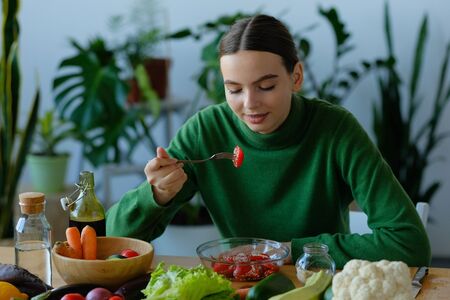Young and happy woman eating healthy salad sitting on the table with green fresh ingredients indoors. Vegan food concept. World vegan dayの写真素材