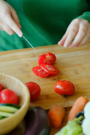 A young girl in a green sweater slices a knife tomatoes on a wooden cutting board to make a salad....
