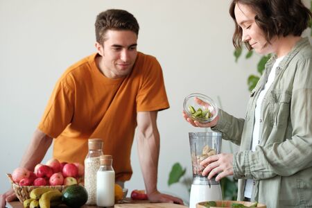 A young married couple is preparing a smoothie: a guy is watching a girl, a girl is filling a kiwi...