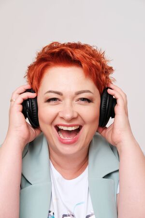 Studio portrait of excited redhead woman listening music in headphones on plain background.The...