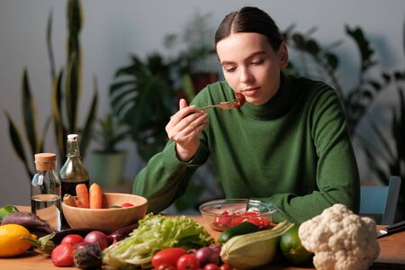 Young and happy woman eating healthy salad sitting on the table with green fresh ingredients...