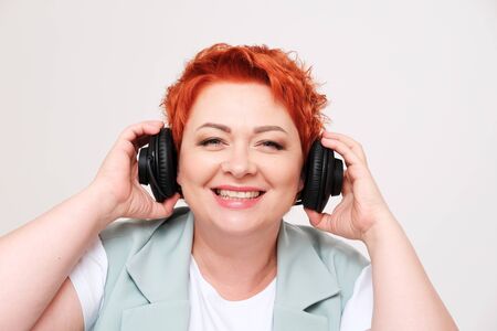 Studio portrait of excited redhead woman listening music in headphones on plain background.The concept of music and radio. Human emotionsの写真素材