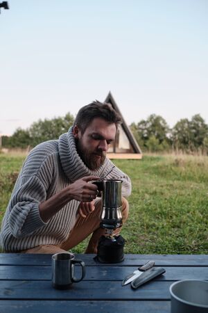 a young man pours coffee from a geyser coffee maker. hipsters in nature. camping conceptの写真素材