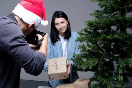 A young guy in a Christmas cap is filming on a mirrorless camera as a young girl with black hair is wrapping Christmas presents. Shooting for blog and vlog. Social Media Videos and Photos. DIY conceptの写真素材