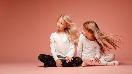 Two cute little girls are sitting next to each other on a pink background in the studio. Hair fluttering in the wind. Kindergarten, childhood, fun, family concept. Two fashionable sisters posing.の写真素材
