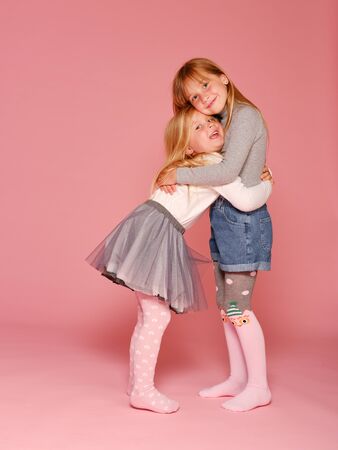 Two cute little girls are standing next to each other on a pink background in the studio. Kindergarten, childhood, fun, family concept. Two fashionable sisters posing.の写真素材