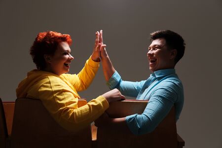Studio portrait of cheerful couple couple gives five to each other on chairs indoors