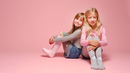 Two cute little girls are sitting next to each other on a pink background in the studio. Kindergarten, childhood, fun, family concept. Two fashionable sisters posing.の写真素材