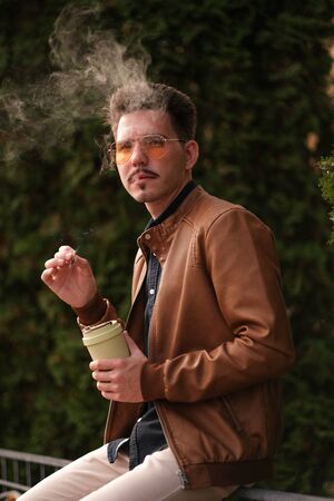 A young guy in yellow sunglasses smokes, releases smoke from his mouth. Harm to health and others....