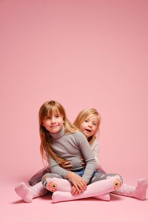 Two cute little girls are sitting next to each other on a pink background in the studio. Kindergarten, childhood, fun, family concept. Two fashionable sisters posing.の写真素材