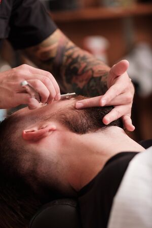 Kazan, Russia, August 19, 2018: Bearded man with long beard, brutal, caucasian hipster getting stylish hair shaving, haircut, with razor by barber or hairdresser with tattoo on hands at barbershopのeditorial素材