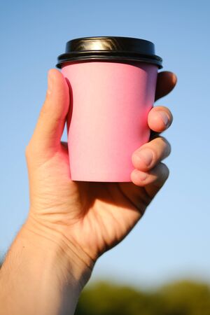 Pink paper coffee cup with a black cap in a male hand against a blue sky. Coffee to go, take-away coffee.の写真素材
