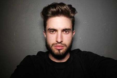 young handsome male with beard, mustache and trendy hairdo, wears casual grey sweater, has serious expression, poses in studio against gray backgroundの写真素材