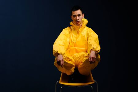 Asian man in a yellow chemical protection suit posing with a yellow plastic chair on a colored trendy background. Fashion shooting in Asian style, night city, neon lights and fashionable clothesの写真素材