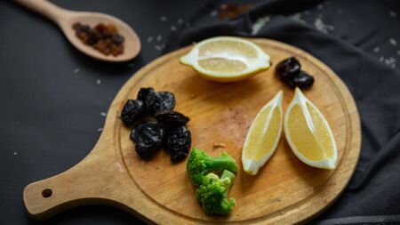 Products on a round wooden board: lemon slices, prunes, broccoli and dried grapes (raisins) in a...
