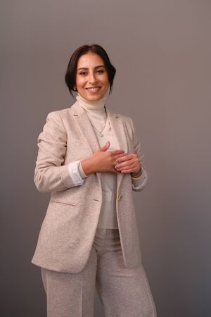 Portrait of a smiling mid-aged business woman (30-35 years old) with dark hair and brown eyes in a milk-colored trouser suit and turtleneck against a light wall. Close-up portrait of a businesswomanの写真素材