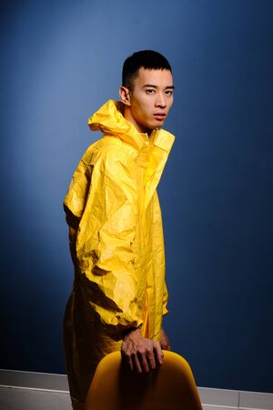 Asian man in a yellow chemical protection suit posing with a yellow plastic chair on a colored trendy background. Fashion shooting in Asian style, night city, neon lights and fashionable clothesの写真素材