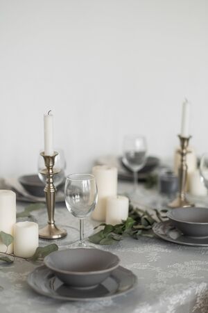 Table setting: gray dishes, white candles, candlesticks, glass goblets, sprigs of eucalyptus on a wooden table with a white tablecloth. Beautiful Scandinavian style interior.の写真素材