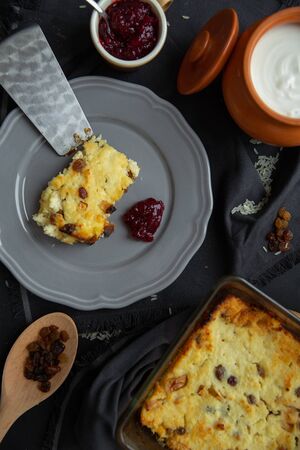 Cottage cheese casserole with raisins on a gray plate, poured sour cream on top. Flat lay, top view. Pot with sour cream, casserole, jar of jam, a wooden spoon with raisins on a dark backgroundの写真素材