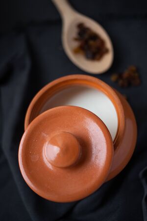Brown ceramic pot with sour cream and a wooden spoon with raisins (dry grapes) on a dark background. Healthy food, dairy products. Farm Products Concept. Top Viewの写真素材