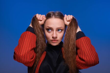 a young girl with blue eyes and blond long hair in a black T-shirt and red bomber jacket on a trendy blue background. Fashion lifestyle portrait of joyful woman, holding her hairs like two ponytailsの写真素材