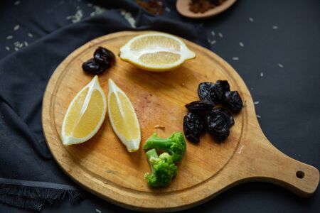 Products on a round wooden board: lemon slices, prunes, broccoli and dried grapes (raisins) in a wooden spoon. Ingredients for cooking.の写真素材
