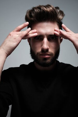 A young dark-haired man with a beard holds his head and shows different human emotions: hatred, fear, despair, horror, malaise, headache, clairvoyance. Close-up studio portrait of a man.の写真素材