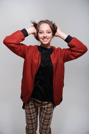 Portrait of a young funny girl with blue eyes and brown hair in a black T-shirt and a red bomber jacket. The girl holds on to her hair and fools around, has fun, grimaces, shows human emotionsの写真素材