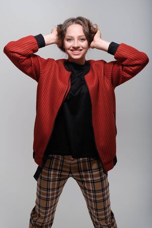 Portrait of a young funny girl with blue eyes and brown hair in a black T-shirt and a red bomber jacket. The girl holds on to her hair and fools around, has fun, grimaces, shows human emotionsの写真素材