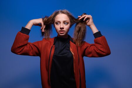 a young girl with blue eyes and blond long hair in a black T-shirt and red bomber jacket on a trendy blue background. Fashion lifestyle portrait of joyful woman, holding her hairs like two ponytailsの写真素材