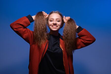 a young girl with blue eyes and blond long hair in a black T-shirt and red bomber jacket on a trendy blue background. Fashion lifestyle portrait of joyful woman, holding her hairs like two ponytailsの写真素材