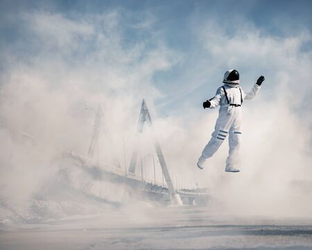 An astronaut in a spacesuit flies in a cloud of smoke against the background of a bridge and urban structures. The astronaut is exploring the planet Earth. Cosmonautics day and earth day concept.の写真素材