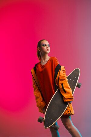 A young girl with bright makeup holds a longboard in her hands on a fashionable neon color background. Portrait of a girl skater. The concept of leisure, extreme sports, healthy lifestyleの写真素材