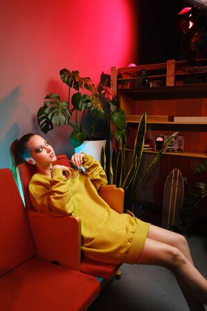 A young attractive girl with bright makeup and in colored clothes sits on a red armchair against a fashionable neon background. The interior of a modern teenager - a lot of flowers and longboardsの写真素材