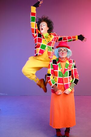 Two clowns a man and a woman with makeup in bright colored costumes are fooling around and showing a presentation using various accessories. April Fools Day concept. Birthday for kidsの写真素材